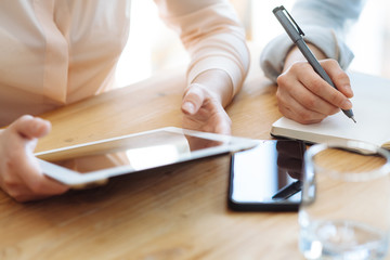 Two female businesswomen working with hands on a tablet in an office in sunlight at a wooden table and one of them writing a pen in a stylish blank on the desk is a modern smartphone