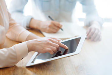 Two female businesswomen working their hands on a tablet in an office in the sunlight at a wooden table and one of them writes a pen in a stylish notebook, and touches the tablet with her finger