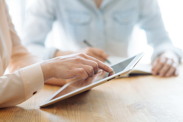 Two female businesswomen working their hands on a tablet in an office in the sunlight at a wooden table and one of them writes a pen in a stylish notebook, and touches the tablet with her finger