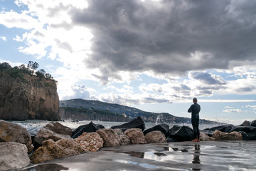 man in a wet suit, surfer, standing on the shore and looking at the waves in the background of the mountain, Sorrento Italy