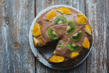 Homemade sponge cake with chocolate cacao ganache frosting. Slices of kiwi and orange on the dessert. Baked pastry in the kitchen on the old vintage wooden background.