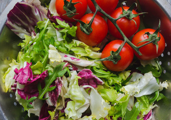 Fresh salad in the steel aluminium colander. Lettuce, arugula and red cherry tomatoes on the vine. Vegan summer food in the kitchen on old vintage wooden background. Vegetarian and healthy vegetables.