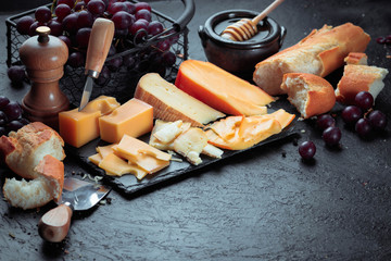 Black slate board with various cheese, decorated with grapes, bread and honey