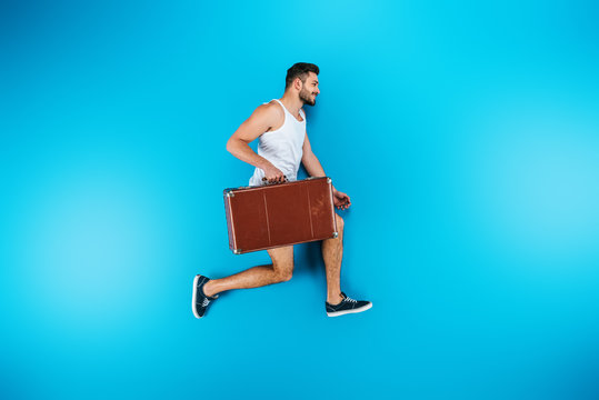 Handsome Smiling Young Man Holding Suitcase And Running On Blue