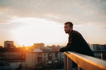 Young man is pondering on terrace of roof against background of cityscape at sunset.