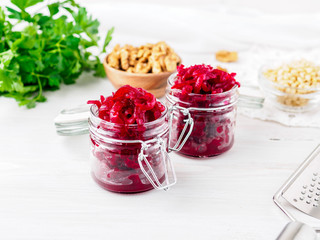 Fresh salad of grated boiled beetroot in glass jars, white wooden background, side view.