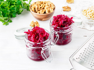 Fresh salad of grated boiled beetroot in glass jars, white wooden background, side view.