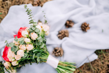 a wedding bouquet of pink roses and peonies lies on a gray fabric with cones on the ground in the woods