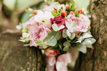 close-up bridal bouquet of roses and peonies with red and pink ribbons lies between trees