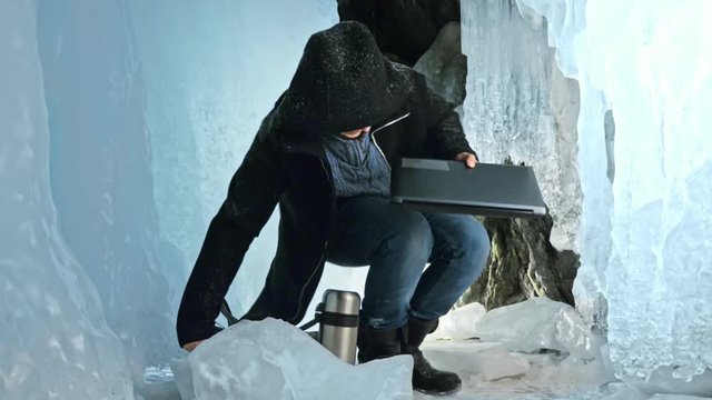 Hacker hacks the server. Man to program on laptop in ice cave. Around the mysterious ice grotto. Man hides himself in a hood. Hacker has criminal cases on the Internet. Guy is warming himself with the