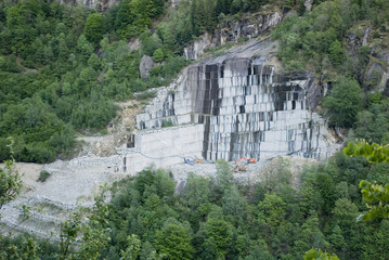 Fototapeta na wymiar landscape: rock quarry of gray granite, in the mountain, forest, rectangular stone blocks carved out of the explosion with dynamite, digger moves rocks to be transported by trucks, Piedmont, Italy