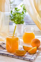 Juicy orange juice in a glass and jug and ripe fruit orange on a white table near the window, the concept of breakfast on a sunny summer day