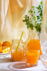 Juicy orange juice in a glass and jug and ripe fruit orange on a white table near the window, the concept of breakfast on a sunny summer day