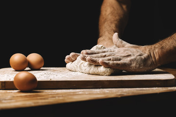 Beautiful and strong men's hands knead the dough from which they will then make bread, pasta or pizza. A cloud of flour flies around like dust. Next to the chicken egg. The background is dark.
