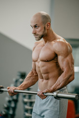 Fototapeta na wymiar Shredded shirtless tough guy with beard and shaved head doing biceps curls with barbell in a gym. Fit man trains.