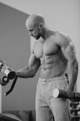 Fototapeta na wymiar Shredded shirtless tough guy with beard and shaved head doing biceps curls with dumbbells in a gym. Fit man trains. Black and white portrait. Black and white portrait.