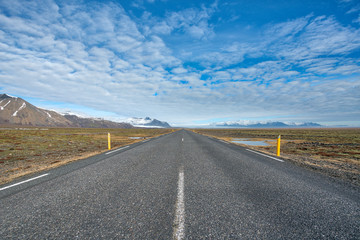 Fototapeta na wymiar Endlessly straight road leading towards glacier and mountains at sunny day with blue half cloudy sky in Iceland