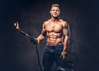 Brutal tattoed male welder with a stylish haircut and beard, with muscular body, dressed in only jeans, holds propane tank and a burning burner, standing in a studio, looking at a camera. - obrazy, fototapety, plakaty