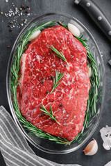 Top view of a raw beef steak for baking in a glass bowl with garlic and rosemary. The concept of a festive table and dishes.