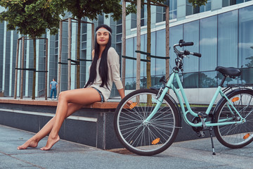 Obraz na płótnie Canvas Portrait of a sexy hippie female wearing blouse and shorts in a headband, sits barefoot on a bench near city bike against a skyscraper