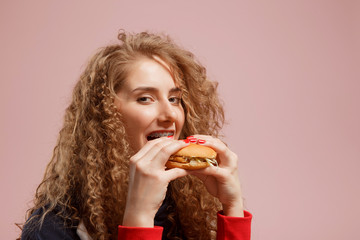 Girl with braces on her teeth bites burger, curly hair, pink background. Concept fast food, clean braces