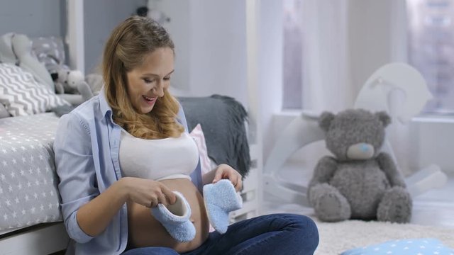Attractive young expecting mother playing with little blue baby shoes on her big naked pregnant belly. Feminine happy woman smiling and talking to unborn baby while stepping on tummy with child shoes
