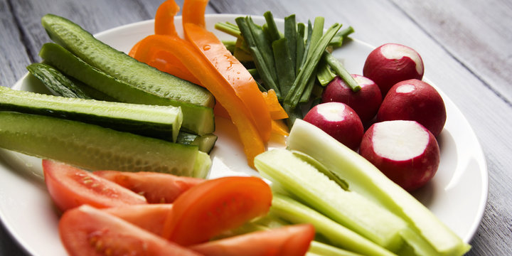 White plate with vegetables for a vegetarian salad. White wooden kitchen table. Radishes, tomatoes, celery, bell pepper, onion and cucumber. Close up. Wide photo.