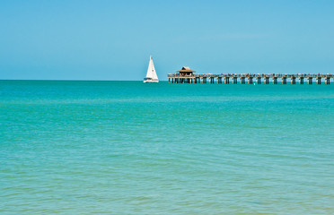 Obraz premium Long view of a sailboat under way, with filled sails passing a wood pier jutting out from a tropical beach with calm waters of the Gulf of Mexico