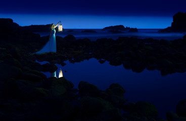 Dark night, blue sea and sky, blonde woman in long dress with lantern