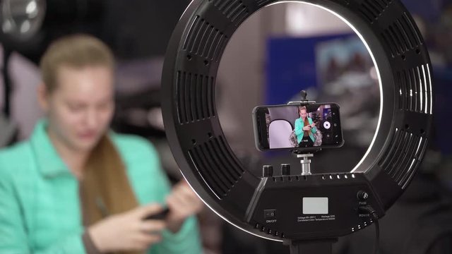 Vlogger recording video blog in front of smartphone. Blogger talking about photographic and video equipment. Young girl uses dimmable LED ring light panel with mobile phone camera mounting adapter.