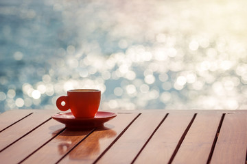 Red coffee cup on wooden table with bright sea on the background.