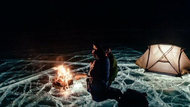 Three travelers by the fire right on the ice at night. Campground on ice. The tent stands next to the fire. People are warming themselves by the fire. Time-lapse with a circular motion. The Lake