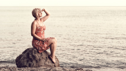 Blonde woman in purple dress sitting on the huge rock next to the sea, looking at the sunset and waiting for something or somebody.