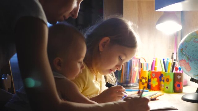 Young mother helps her daughter with her homework at on the table under the light of a lamp. Mom holds a small son in his arms.
