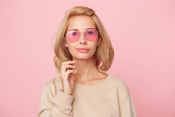 Young pretty, cute woman, touching her hair, standing over pink background, feels happy and joy. Wears yellow sweater and glasses.