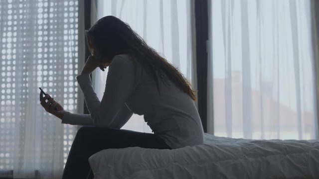 Side shot of a frustrated woman sitting on bed with a big bright window in background.
