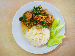 Top view Thai local traditional stir fried spicy holy basil with ground pork, crispy black preserved Century Egg (Kai Yieow Ma), white boiled Jasmine rice, fresh cucumber, on blue plate, creme table