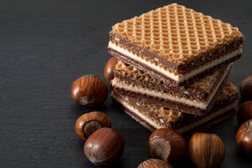 Wafers, cookies and sweets concept with hazelnut, chocolate and vanilla filled wafer stack with raw hazelnuts still in shell scatered around on a dark background