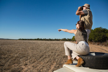 Woman with man pointing while sitting on tire over vehicle hood at field