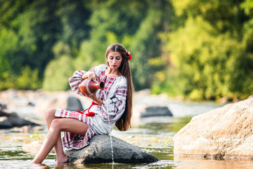 girl in embroidered dress sits on a stone with a jug near the water