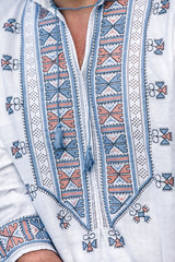 a man stands in a white embroidered shirt with an ornament of blue and red thread