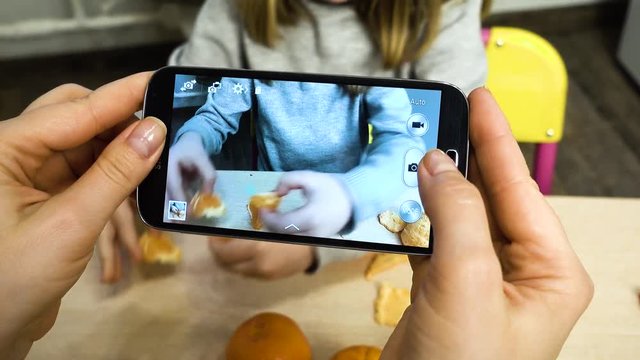 Mom takes pictures on a smartphone, as a child cleans, peeling and eats a mandarin. A girl 5-6 years old is sitting at a children's table.