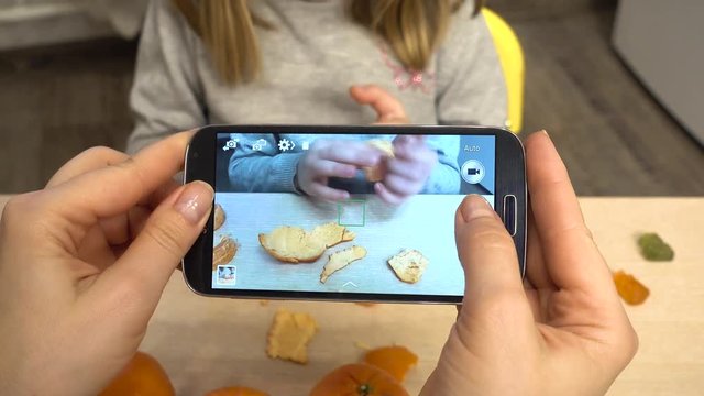 Mom removes the child on the smartphone. A girl 5-6 years old is going to eat a mandarin sitting at a table.