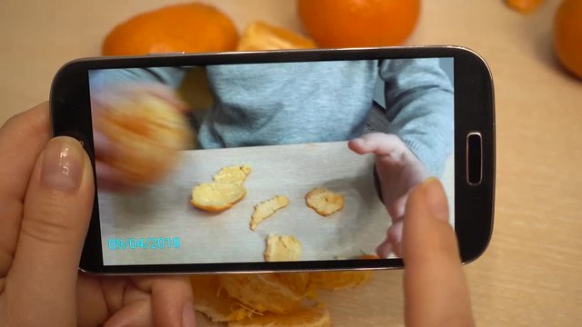 Woman's hand, mother takes pictures on a smartphone, as a child cleans, peeling and eats a mandarin. A girl 5-6 years old is sitting at a children's table. Mom scrolling photos.