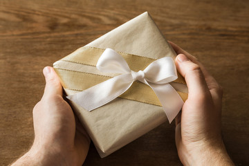 Man's hands holding a gift box with white ribbon on a brown wooden desk. Giving or receiving a lovely gift. Congratulation concept. 