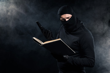 Man in black balaclava holding gun and reading book