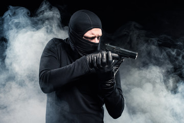 Robber in balaclava and gloves aiming with gun in clouds of smoke