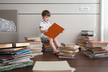 The boy sits on books and reads. Around him are many colorful books.