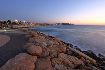 Promenada nadmorka w Tel Awiwie, Izrael, kamienie umacniające brzeg, morze, światła latarń ulicznych, w oddali, na horyzoncie, widoczna Hajfa, wieczór, długie naświetlanie