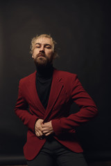 Modern businessman. Handsome young beautiful and confident bearded blond man, wearing black shirt and red jacket, standing in studio with dark background